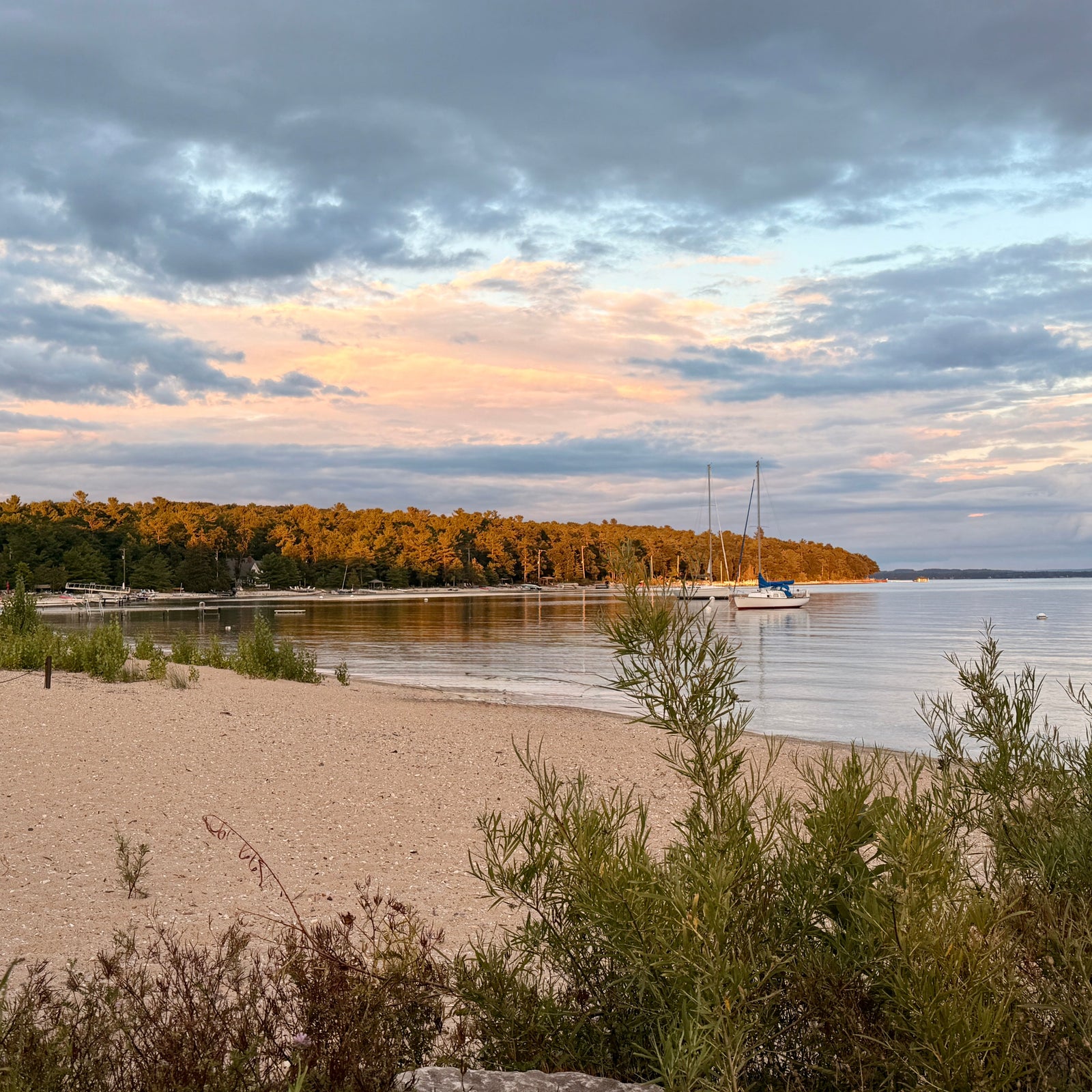 A scenic view of Haserot Beach and Old Mission Harbor under a cloudy sky at sunset, with a line of trees showing autumn colors in the distance and a few boats on the water.